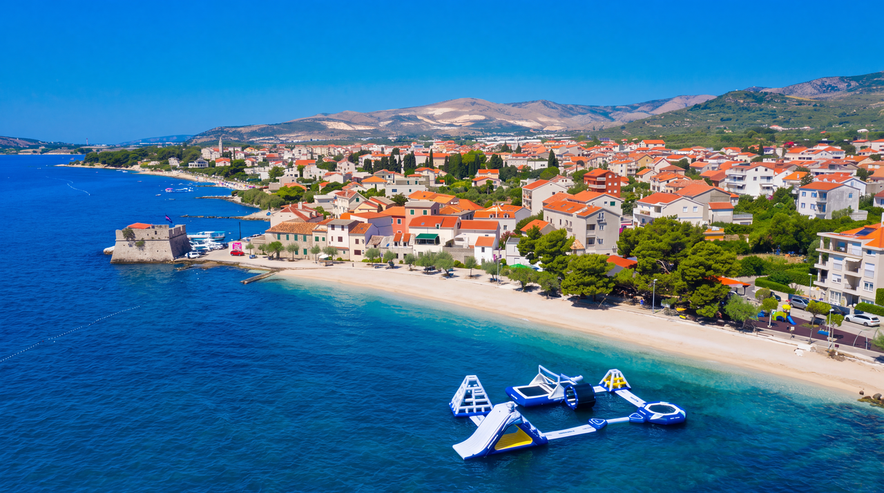 The Kaštela coast from the air: a pale beach curving along clear turquoise water, red-roofed houses behind it and the hills rising beyond.