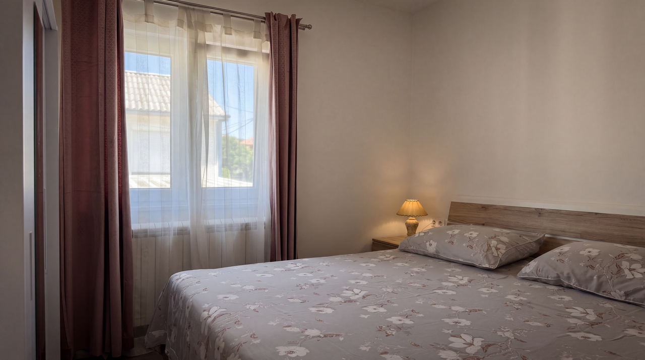The bedroom: a double bed in floral linen against a timber headboard, a small shaded lamp beside it, and a curtained window letting in the morning light.