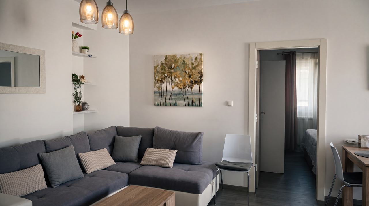 The living room: a grey corner sofa under a framed painting of trees, three glass pendant lights over a timber table, and an open door through to the bedroom.