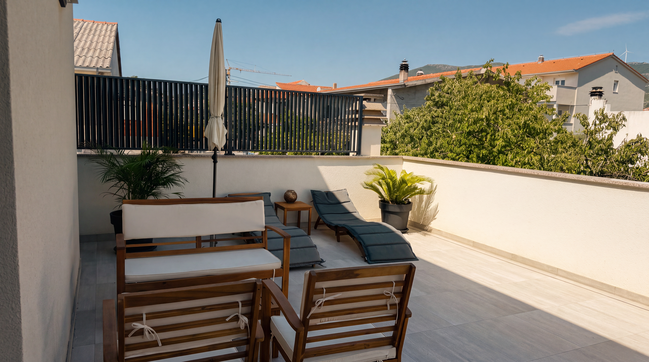 The terrace in full sun: teak chairs and a cushioned bench, a closed parasol and a sun lounger along the rail, with tiled roofs and green hills past the balustrade.